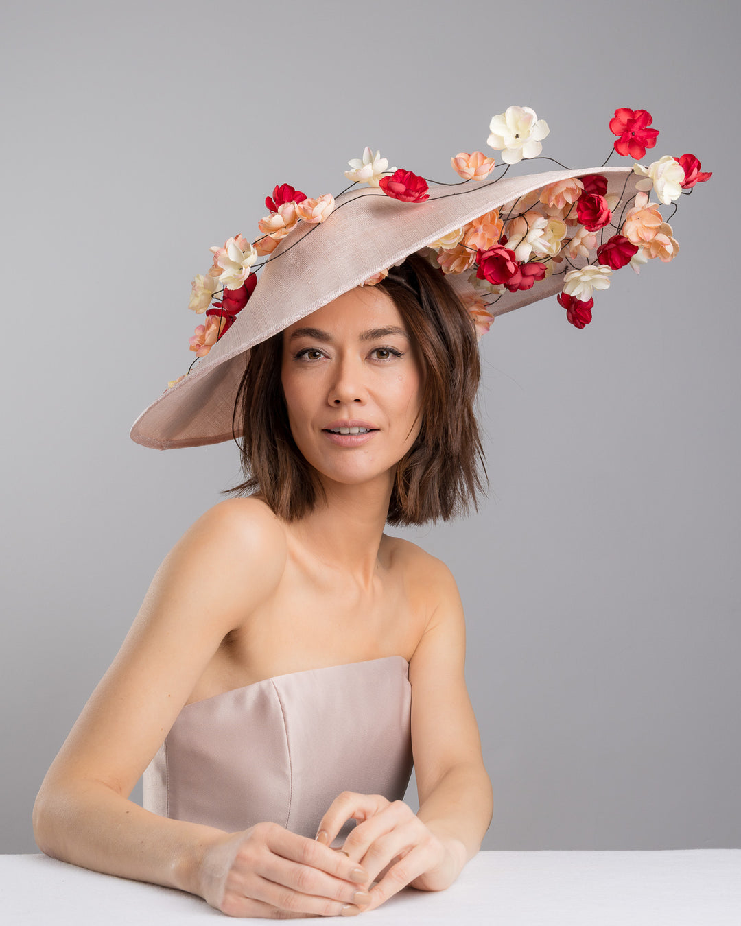 The female model is posing, wearing
Rivane, the soft blush sinamay base hat with an elegant sculpted brim, adorned with cascading clusters of cream, peach, and deep red flowers on fine wired stems, creating a romantic, floating garden effect. Designed and made by Merve Bayindir Millinery.

