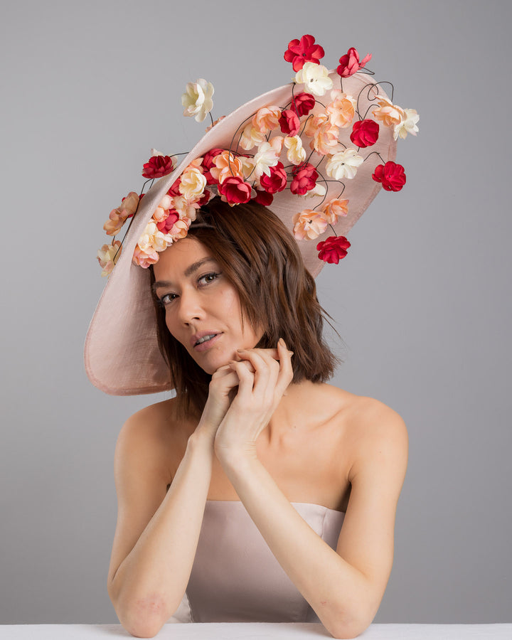 The female model is posing, wearing
Rivane, the soft blush sinamay base hat with an elegant sculpted brim, adorned with cascading clusters of cream, peach, and deep red flowers on fine wired stems, creating a romantic, floating garden effect. Designed and made by Merve Bayindir Millinery.

