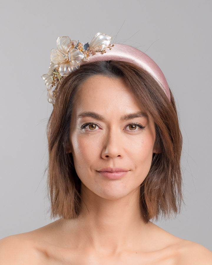 The model is showcasing the front side close up of the blush pink satin padded headband featuring sculptural translucent ivory flowers with fine gold edging. The design is accented with delicate metallic leaves, soft blush crystal details, and subtle beadwork arranged asymmetrically along one side, creating an elegant and luminous statement piece suitable for weddings and formal occasions. Designed and made by Merve Bayindir Millinery.
