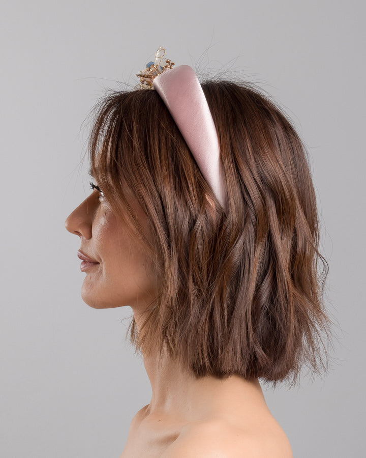 The model is showcasing the left side close up of the blush pink satin padded headband featuring sculptural translucent ivory flowers with fine gold edging. The design is accented with delicate metallic leaves, soft blush crystal details, and subtle beadwork arranged asymmetrically along one side, creating an elegant and luminous statement piece suitable for weddings and formal occasions. Designed and made by Merve Bayindir Millinery.
