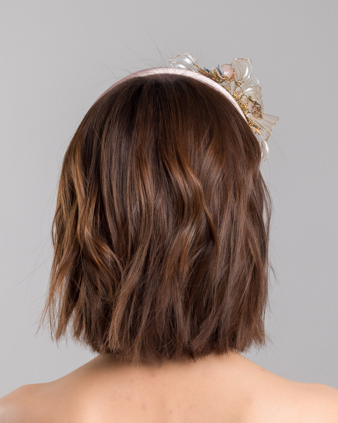 The model is showcasing the back side close up of the blush pink satin padded headband featuring sculptural translucent ivory flowers with fine gold edging. The design is accented with delicate metallic leaves, soft blush crystal details, and subtle beadwork arranged asymmetrically along one side, creating an elegant and luminous statement piece suitable for weddings and formal occasions. Designed and made by Merve Bayindir Millinery.
