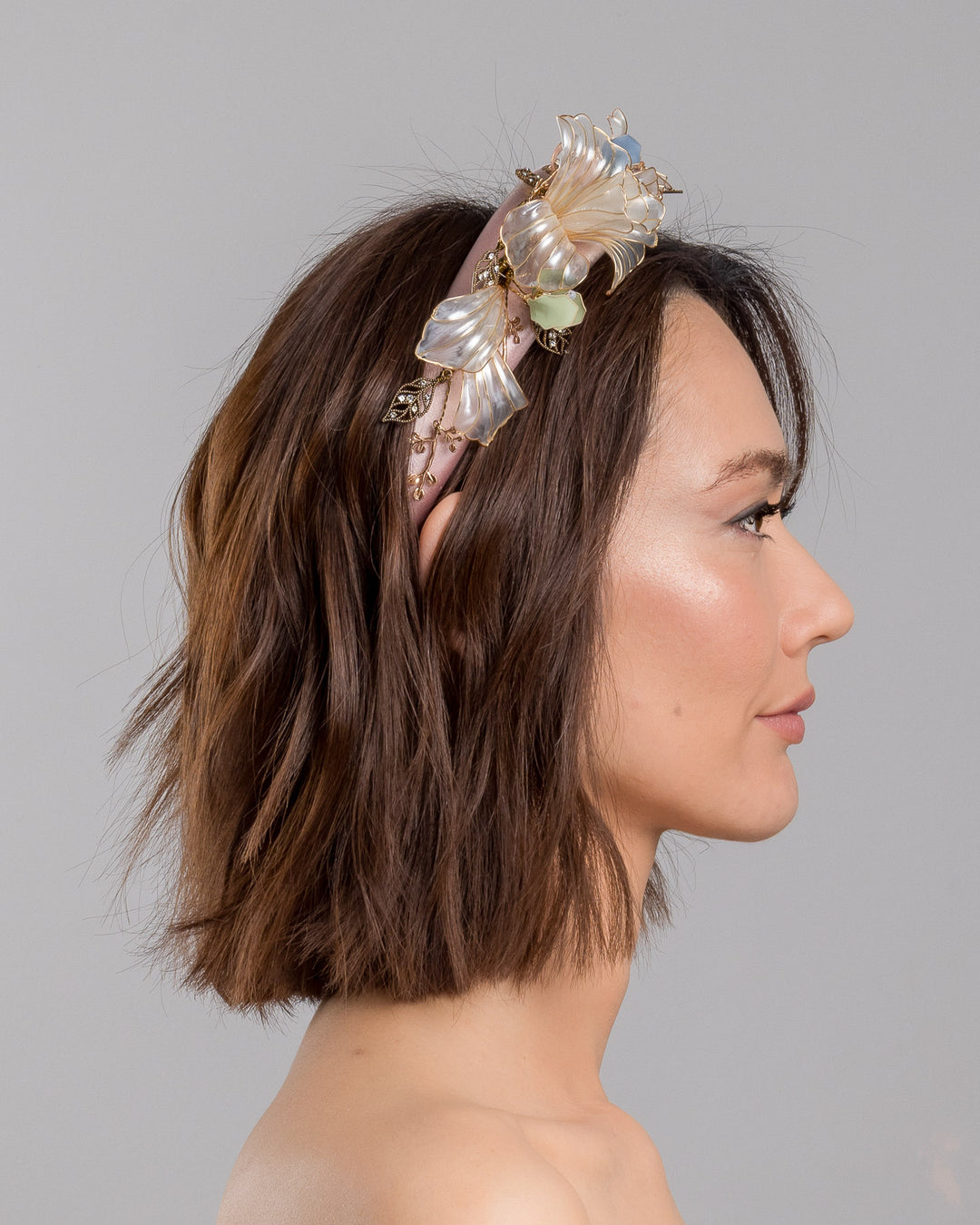 The model is showcasing the right side close up of the blush pink satin padded headband featuring sculptural translucent ivory flowers with fine gold edging. The design is accented with delicate metallic leaves, soft blush crystal details, and subtle beadwork arranged asymmetrically along one side, creating an elegant and luminous statement piece suitable for weddings and formal occasions. Designed and made by Merve Bayindir Millinery.
