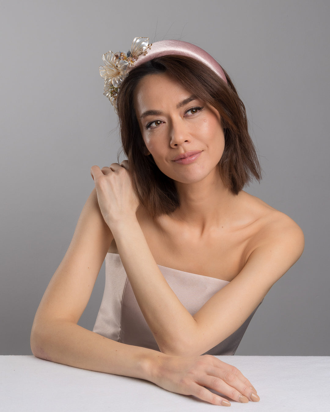 The female model is posing, wearing Rosel, the blush pink satin padded headband featuring sculptural translucent ivory flowers with fine gold edging. The design is accented with delicate metallic leaves, soft blush crystal details, and subtle beadwork arranged asymmetrically along one side, creating an elegant and luminous statement piece suitable for weddings and formal occasions. Designed and made by Merve Bayindir Millinery.
