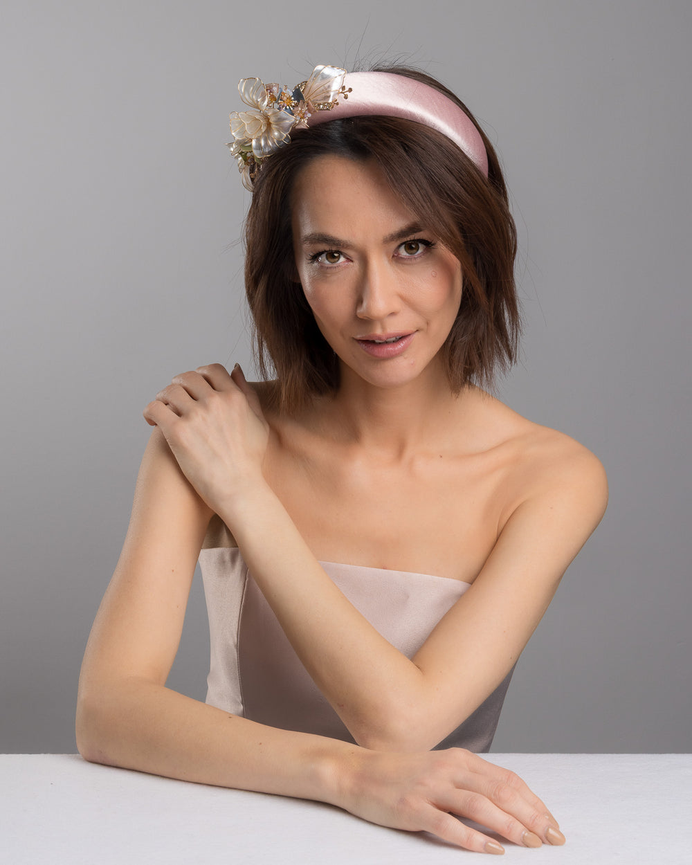 The female model is posing, wearing Rosel, the blush pink satin padded headband featuring sculptural translucent ivory flowers with fine gold edging. The design is accented with delicate metallic leaves, soft blush crystal details, and subtle beadwork arranged asymmetrically along one side, creating an elegant and luminous statement piece suitable for weddings and formal occasions. Designed and made by Merve Bayindir Millinery.
