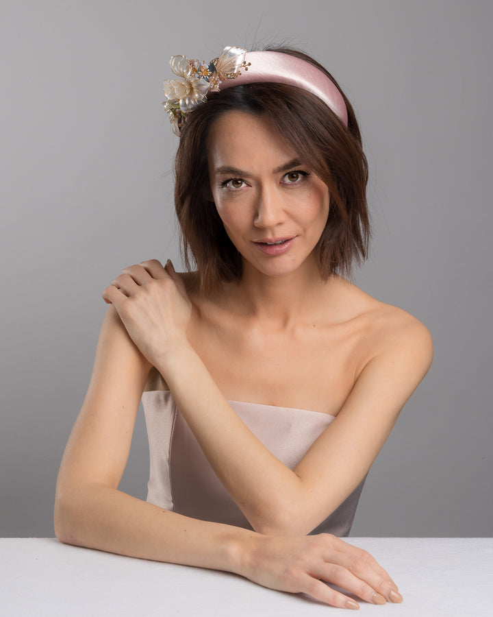 The female model is posing, wearing Rosel, the blush pink satin padded headband featuring sculptural translucent ivory flowers with fine gold edging. The design is accented with delicate metallic leaves, soft blush crystal details, and subtle beadwork arranged asymmetrically along one side, creating an elegant and luminous statement piece suitable for weddings and formal occasions. Designed and made by Merve Bayindir Millinery.
