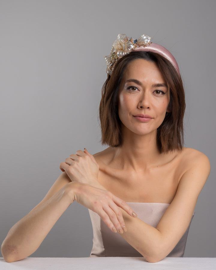 The female model is posing, wearing Rosel, the blush pink satin padded headband featuring sculptural translucent ivory flowers with fine gold edging. The design is accented with delicate metallic leaves, soft blush crystal details, and subtle beadwork arranged asymmetrically along one side, creating an elegant and luminous statement piece suitable for weddings and formal occasions. Designed and made by Merve Bayindir Millinery.
