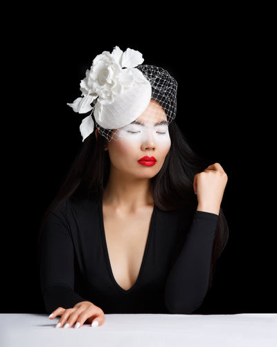 Model is showcasing a White mat sateen covered button shaped fascinator decorated with custom wild crepe white rose that takes the back half of the hat. Finished with a birdcage veil.