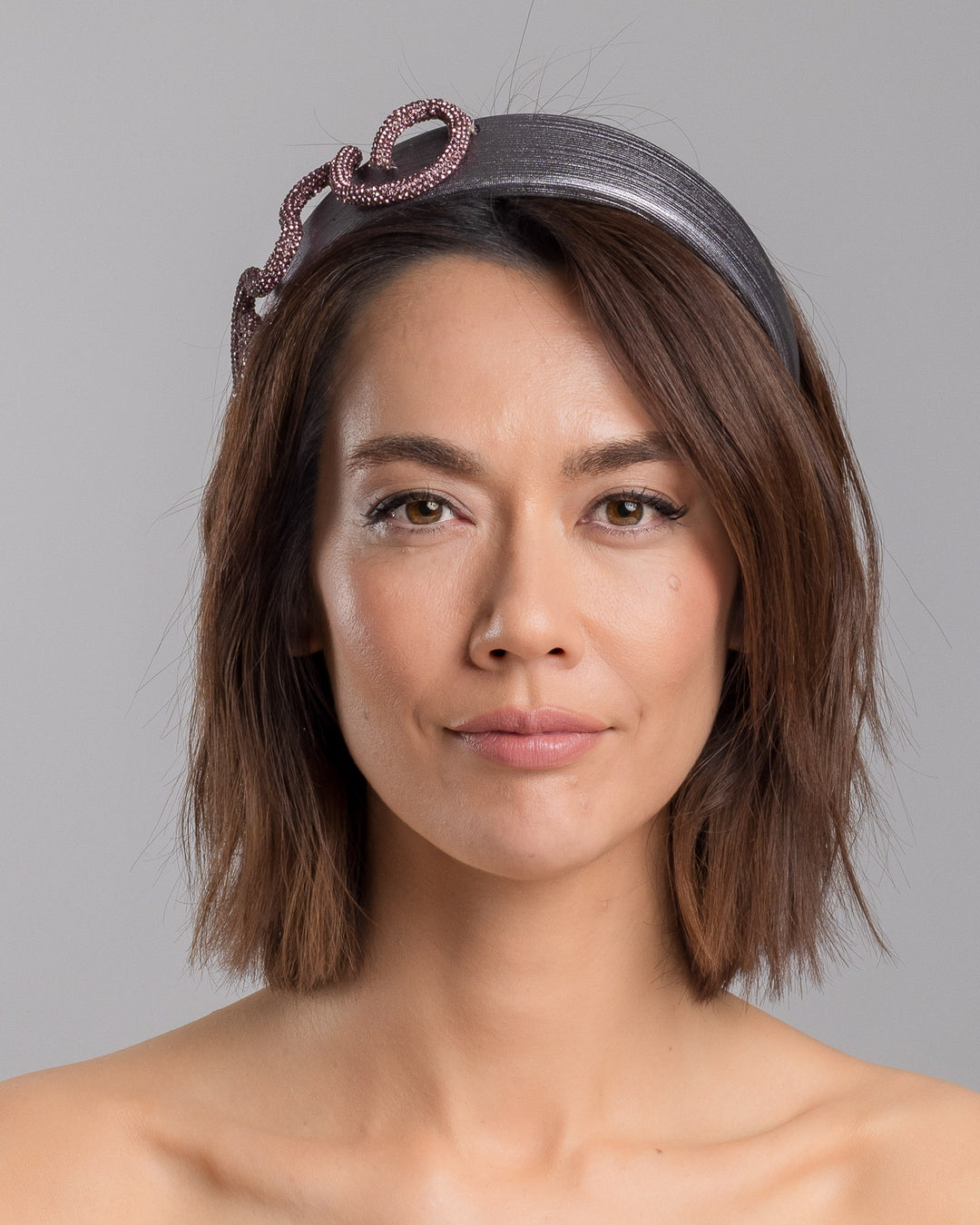 The model is showcasing the front side close up of the charcoal silver headband with a wide faux leather base, adorned with a sculptural wave of rose-pink crystal-embellished cord looping across the top in an elegant, asymmetrical design. Designed and made by Merve Bayindir Millinery.
