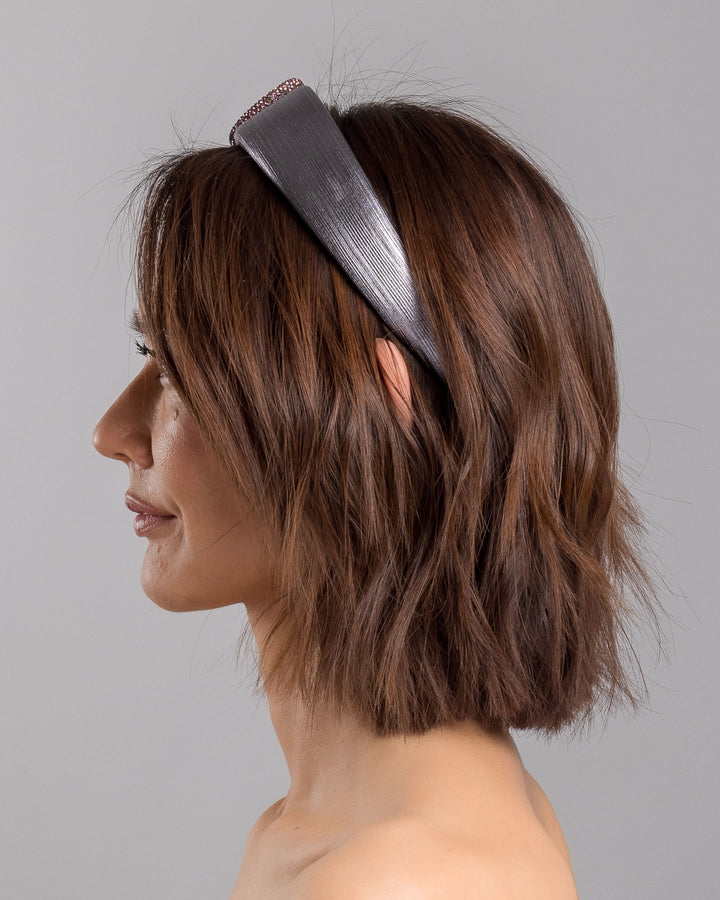 The model is showcasing the left side close up of the charcoal silver headband with a wide faux leather base, adorned with a sculptural wave of rose-pink crystal-embellished cord looping across the top in an elegant, asymmetrical design. Designed and made by Merve Bayindir Millinery.
