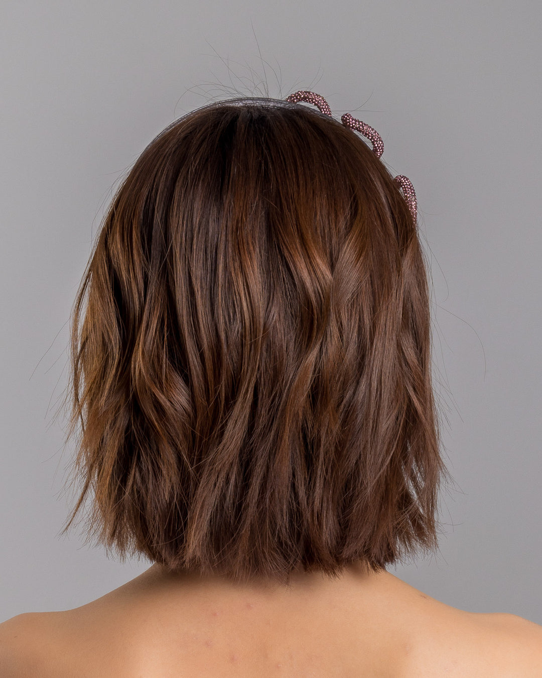 The model is showcasing the back side close up of the charcoal silver headband with a wide faux leather base, adorned with a sculptural wave of rose-pink crystal-embellished cord looping across the top in an elegant, asymmetrical design. Designed and made by Merve Bayindir Millinery.
