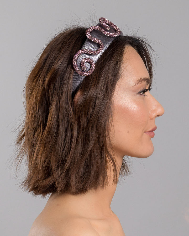 The model is showcasing the right side close up of the charcoal silver headband with a wide faux leather base, adorned with a sculptural wave of rose-pink crystal-embellished cord looping across the top in an elegant, asymmetrical design. Designed and made by Merve Bayindir Millinery.
