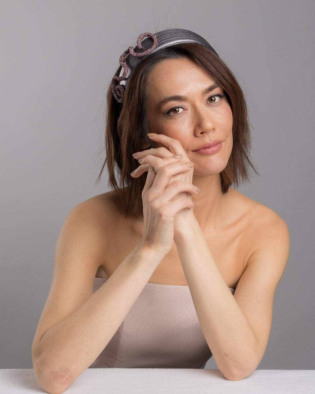 The female model is posing, wearing Rouxel, the charcoal silver headband with a wide faux leather base, adorned with a sculptural wave of rose-pink crystal-embellished cord looping across the top in an elegant, asymmetrical design. Designed and made by Merve Bayindir Millinery.
