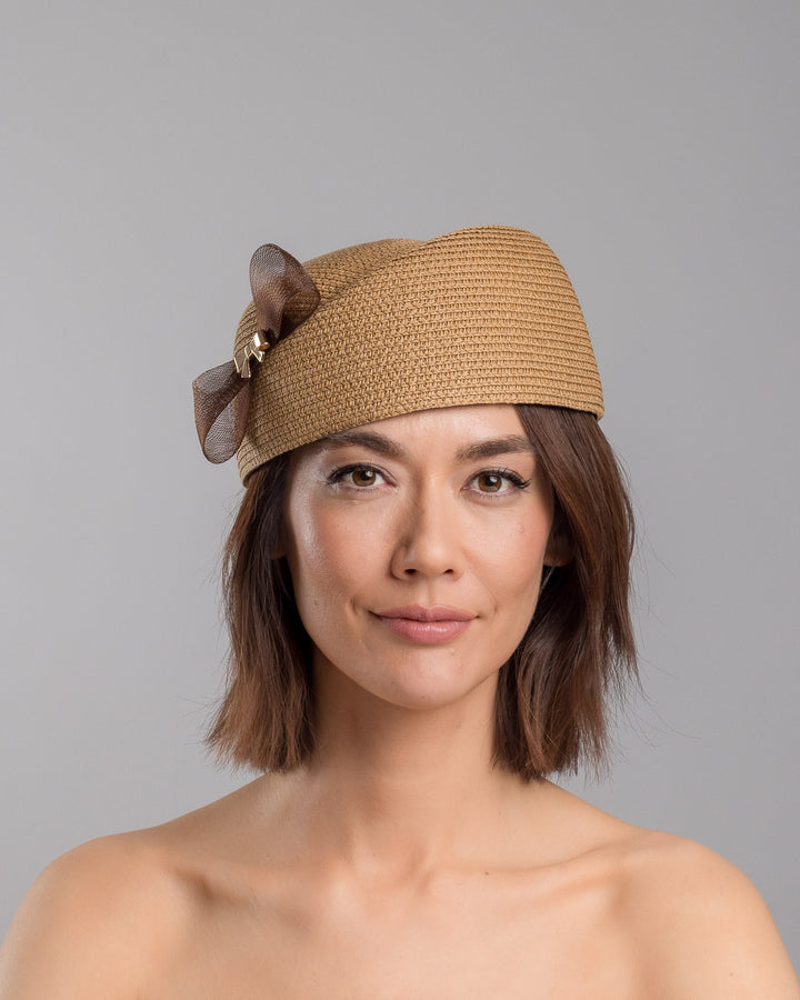 The model is showcasing the front side close up of the natural straw pillbox hat with a structured woven crown, accented by a dark brown mesh bow and a small metallic geometric embellishment at the center for a refined, modern touch. Designed and made by Merve Bayindir Millinery.
