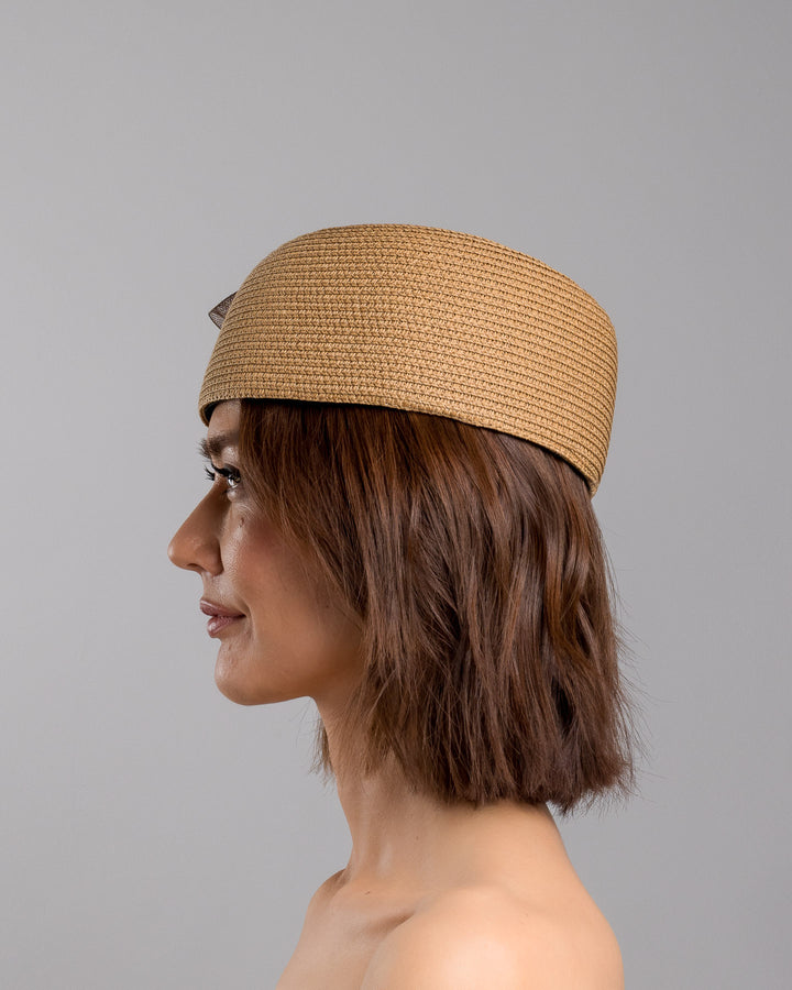 The model is showcasing the left side close up of the natural straw pillbox hat with a structured woven crown, accented by a dark brown mesh bow and a small metallic geometric embellishment at the center for a refined, modern touch. Designed and made by Merve Bayindir Millinery.
