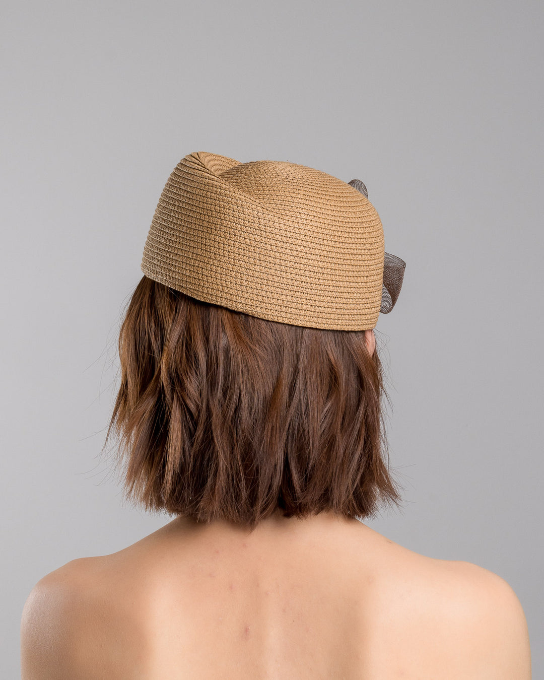 The model is showcasing the back side close up of the natural straw pillbox hat with a structured woven crown, accented by a dark brown mesh bow and a small metallic geometric embellishment at the center for a refined, modern touch. Designed and made by Merve Bayindir Millinery.
