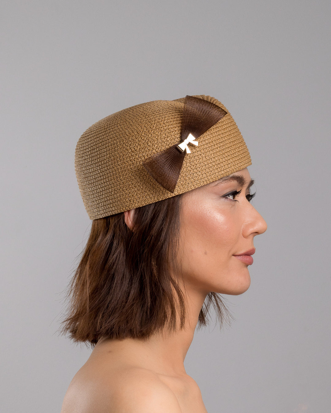The model is showcasing the right side close up of the natural straw pillbox hat with a structured woven crown, accented by a dark brown mesh bow and a small metallic geometric embellishment at the center for a refined, modern touch. Designed and made by Merve Bayindir Millinery.
