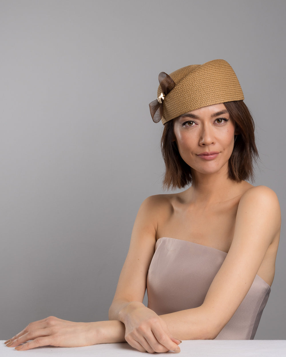 The female model is posing, wearing
Rye, the natural straw pillbox hat with a structured woven crown, accented by a dark brown mesh bow and a small metallic geometric embellishment at the center for a refined, modern touch. Designed and made by Merve Bayindir Millinery.
