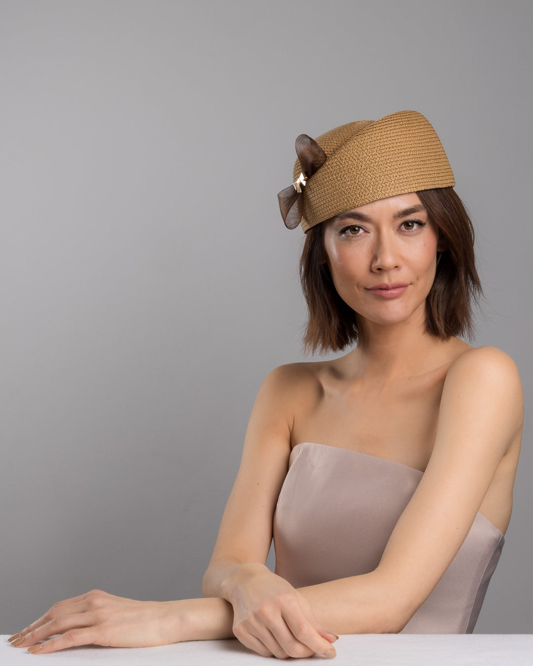 The female model is posing, wearing
Rye, the natural straw pillbox hat with a structured woven crown, accented by a dark brown mesh bow and a small metallic geometric embellishment at the center for a refined, modern touch. Designed and made by Merve Bayindir Millinery.
