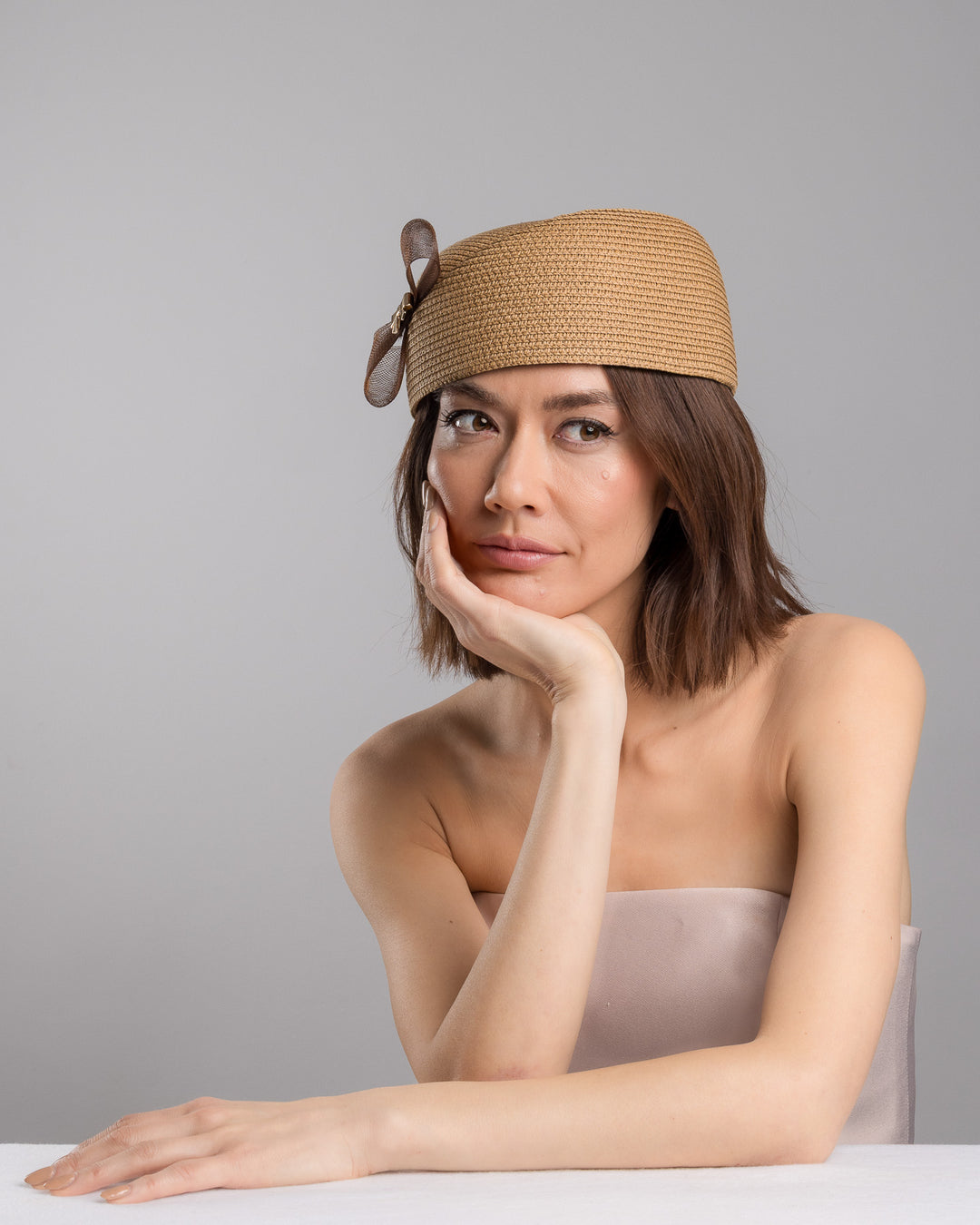 The female model is posing, wearing
Rye, the natural straw pillbox hat with a structured woven crown, accented by a dark brown mesh bow and a small metallic geometric embellishment at the center for a refined, modern touch. Designed and made by Merve Bayindir Millinery.
