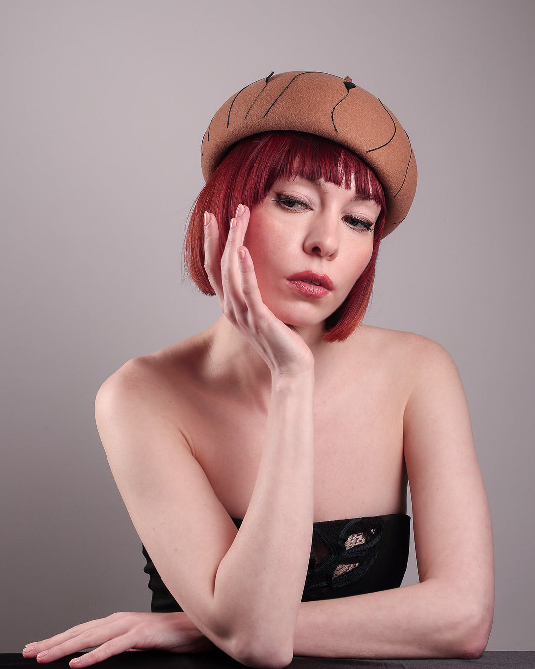 The model showcases theright front of the Raven pillbox beret, crafted in warm caramel felt. Its sculptural silhouette is softened by delicate, trimmed black feather appliqué lines cascading around the hat. Designed by Merve Bayindir.
