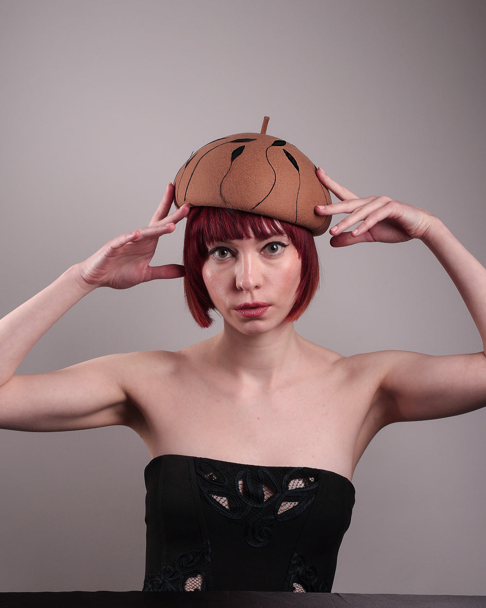 The model showcases the front of the Raven pillbox beret, crafted in warm caramel felt. Its sculptural silhouette is softened by delicate, trimmed black feather appliqué lines cascading around the hat. Designed by Merve Bayindir.