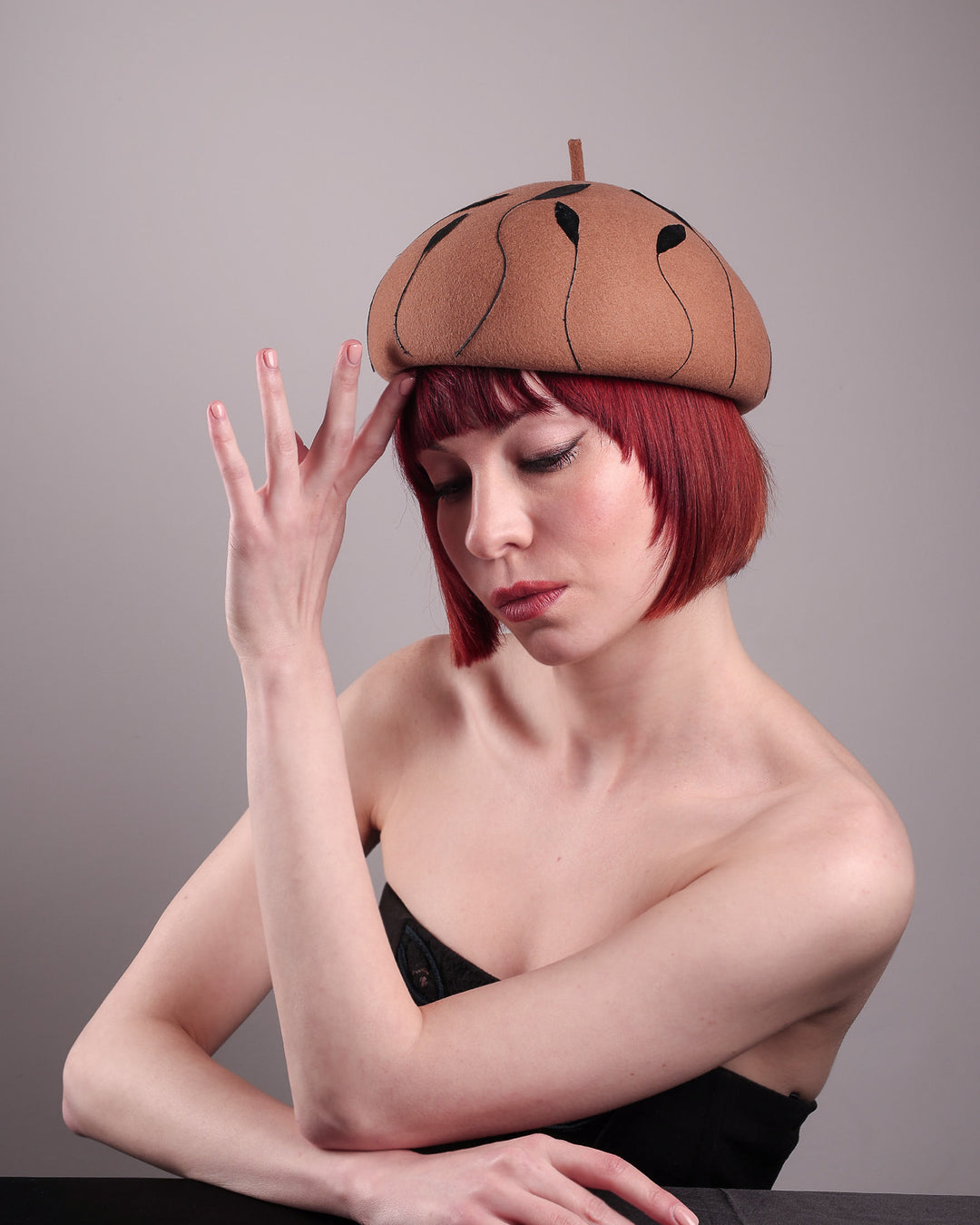 The model showcases the right front of the Raven pillbox beret, crafted in warm caramel felt. Its sculptural silhouette is softened by delicate, trimmed black feather appliqué lines cascading around the hat. Designed by Merve Bayindir.
