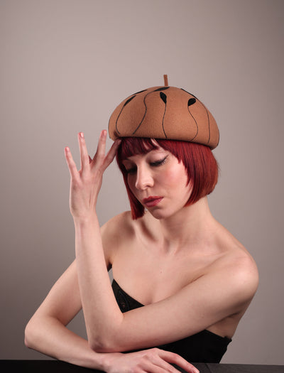 The model showcases the right front of the Raven pillbox beret, crafted in warm caramel felt. Its sculptural silhouette is softened by delicate, trimmed black feather appliqué lines cascading around the hat. Designed by Merve Bayindir.
