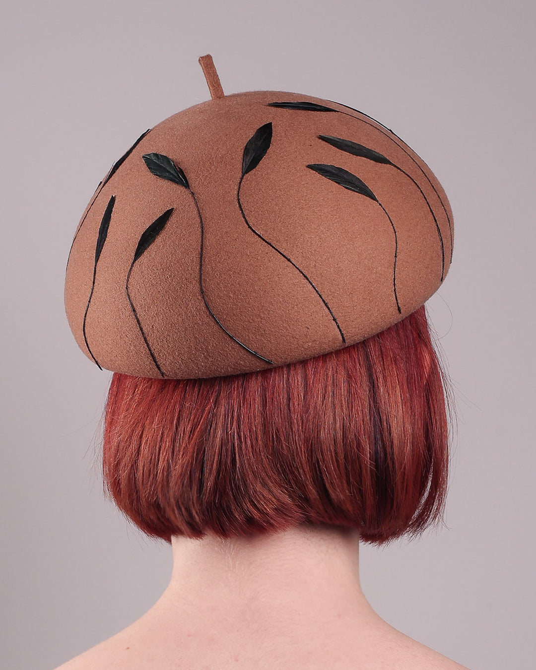 The model showcases the back of the Raven pillbox beret, crafted in warm caramel felt. Its sculptural silhouette is softened by delicate, trimmed black feather appliqué lines cascading around the hat. Designed by Merve Bayindir.