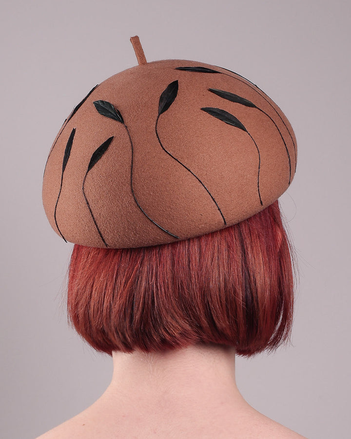 The model showcases the back of the Raven pillbox beret, crafted in warm caramel felt. Its sculptural silhouette is softened by delicate, trimmed black feather appliqué lines cascading around the hat. Designed by Merve Bayindir.