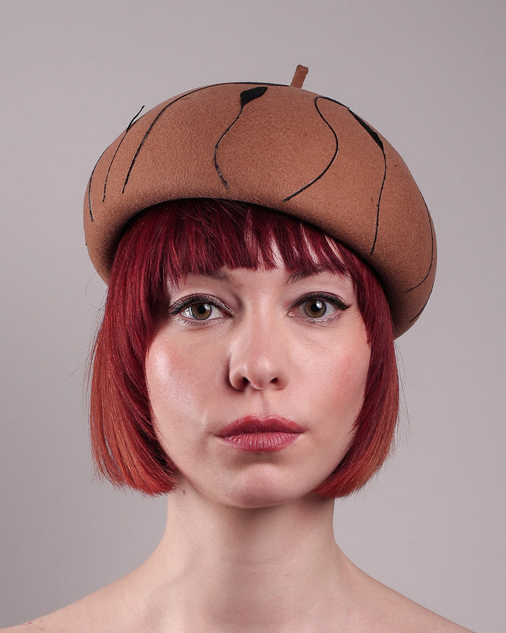 The model showcases the front close up of the Raven pillbox beret, crafted in warm caramel felt. Its sculptural silhouette is softened by delicate, trimmed black feather appliqué lines cascading around the hat. Designed by Merve Bayindir.