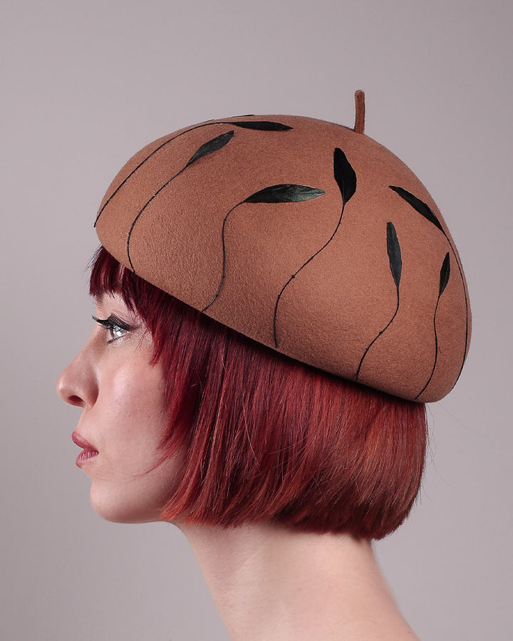 The model showcases the left side close up of the Raven pillbox beret, crafted in warm caramel felt. Its sculptural silhouette is softened by delicate, trimmed black feather appliqué lines cascading around the hat. Designed by Merve Bayindir.