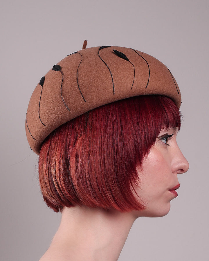 The model showcases the right side close up of the Raven pillbox beret, crafted in warm caramel felt. Its sculptural silhouette is softened by delicate, trimmed black feather appliqué lines cascading around the hat. Designed by Merve Bayindir.