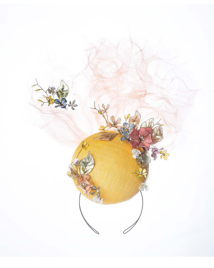 Still life picture of double button yellow fascinator decorated with multi colour  wired resin flowers and peach crinoline clouds.