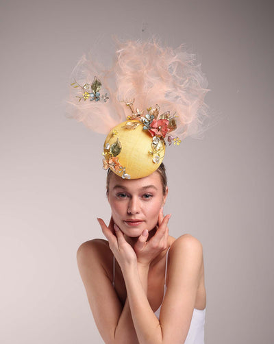 Model is showcasing a double button yellow fascinator decorated with multi colour wired resin flowers and peach crinoline clouds. The fascinator sits in the front centre of the head.