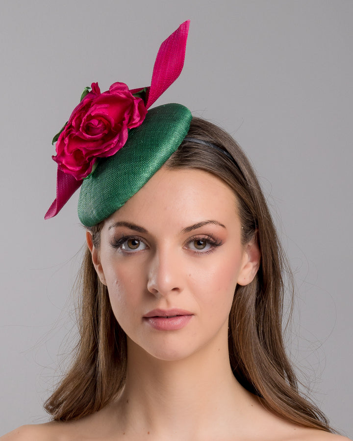 Model is showcasing the front side close up of button-shaped green fascinator with a pink jinsin swirl crossing the base, the centre of the swirls adorned with a hot pink rose. The fascinator sits on the right front side of the head. Designed and made by Merve Bayindir millinery.