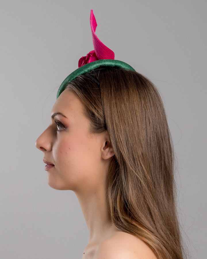 Model is showcasing the left side close up of button-shaped green fascinator with a pink jinsin swirl crossing the base, the centre of the swirls adorned with a hot pink rose. The fascinator sits on the right front side of the head. Designed and made by Merve Bayindir millinery.