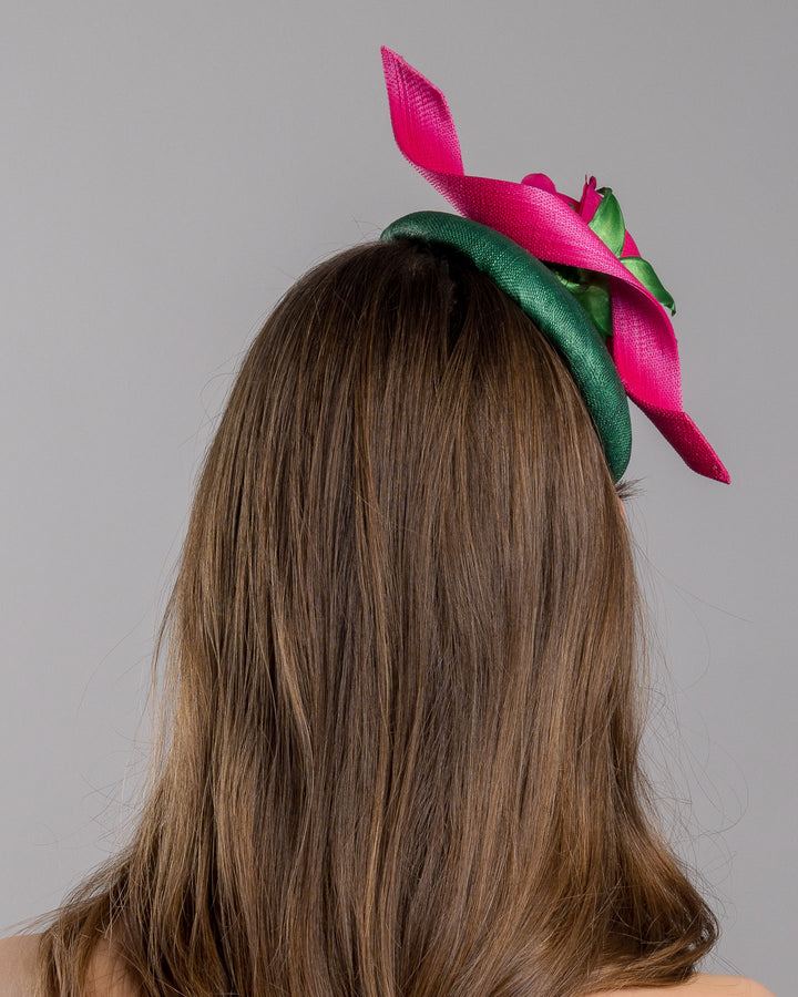 Model is showcasing the back side close up of button-shaped green fascinator with a pink jinsin swirl crossing the base, the centre of the swirls adorned with a hot pink rose. The fascinator sits on the right front side of the head. Designed and made by Merve Bayindir millinery.