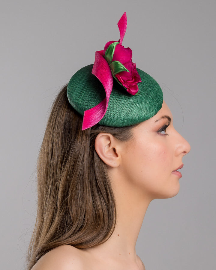 Model is showcasing the right side close up of button-shaped green fascinator with a pink jinsin swirl crossing the base, the centre of the swirls adorned with a hot pink rose. The fascinator sits on the right front side of the head. Designed and made by Merve Bayindir millinery.