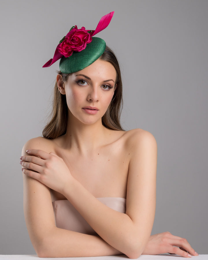 Model is posing wearing the Revel green fascinator with a pink jinsin swirl crossing the base, the centre of the swirls adorned with a hot pink rose. The fascinator sits on the right front side of the head. Designed and made by Merve Bayindir millinery.