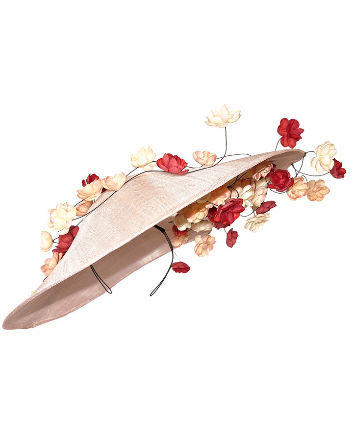 Soft blush sinamay base hat with an elegant sculpted brim, adorned with cascading clusters of cream, peach, and deep red flowers on fine wired stems, creating a romantic, floating garden effect. Designed and made by Merve Bayindir Millinery.
