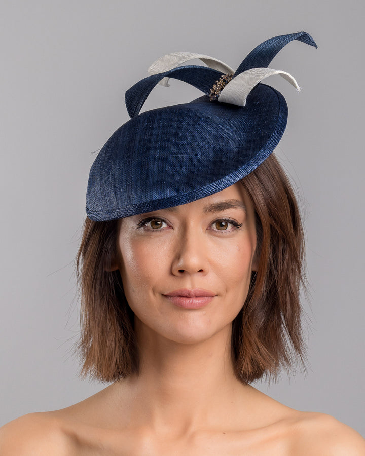 Model is showcasing the front side close up of asymmetric navy saucer, decorated with white and navy buntal swirls and completed with multi coloured crystal beads. The height gained on the left side of this headpiece creates a dramatic effect and completes your style with a unique touch. Designed and made by Merve Bayindir millinery.