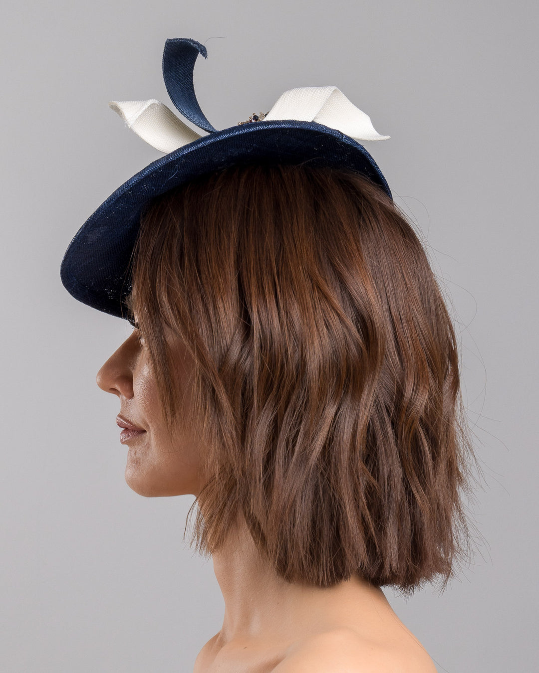 Model is showcasing the left side close up of asymmetric navy saucer, decorated with white and navy buntal swirls and completed with multi coloured crystal beads. The height gained on the left side of this headpiece creates a dramatic effect and completes your style with a unique touch. Designed and made by Merve Bayindir millinery.