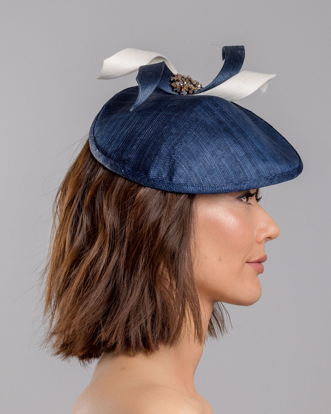 Model is showcasing the right side close up of asymmetric navy saucer, decorated with white and navy buntal swirls and completed with multi coloured crystal beads. The height gained on the left side of this headpiece creates a dramatic effect and completes your style with a unique touch. Designed and made by Merve Bayindir millinery.