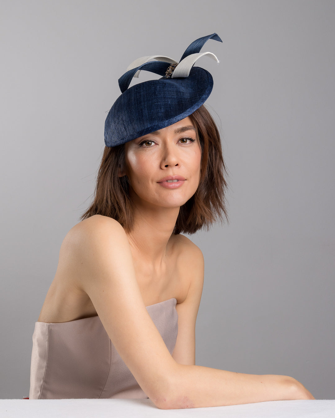 Model is posing wearing Roan, asymmetric navy saucer, decorated with white and navy buntal swirls and completed with multi coloured crystal beads. The height gained on the left side of this headpiece creates a dramatic effect and completes your style with a unique touch. Designed and made by Merve Bayindir millinery.