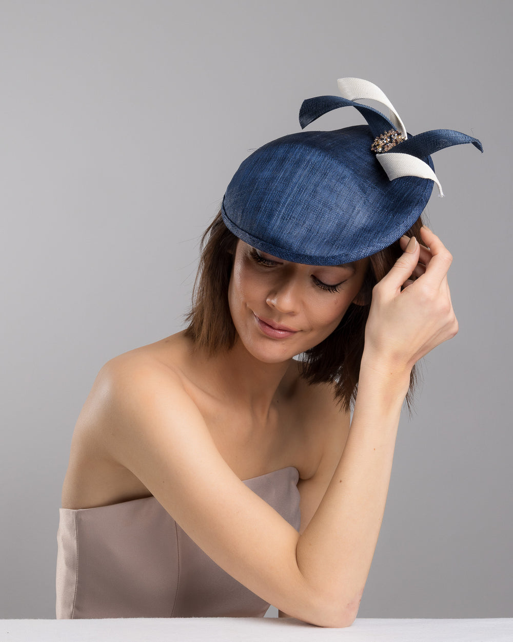 Model is posing wearing Roan, asymmetric navy saucer, decorated with white and navy buntal swirls and completed with multi coloured crystal beads. The height gained on the left side of this headpiece creates a dramatic effect and completes your style with a unique touch. Designed and made by Merve Bayindir millinery.