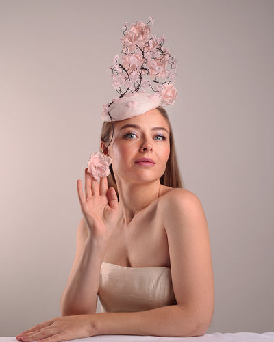 Model is showcasin the right front side of the button shaped pink fascinator with sinamay base decorated with tree like wire branches embellisehd with custom made pink silk flowers. The pink button fascinator is placed on the right front side of the face and the wire braces go up towards the left side of the button base while a single branch with one rose falls on the right side of the face.