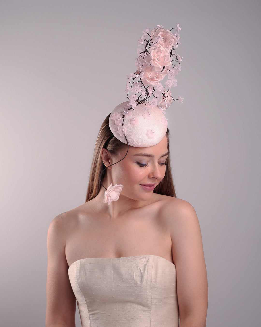 Model is showcasin the right angle of the button shaped pink fascinator with sinamay base decorated with tree like wire branches embellisehd with custom made pink silk flowers. The pink button fascinator is placed on the right front side of the face and the wire braces go up towards the left side of the button base while a single branch with one rose falls on the right side of the face.