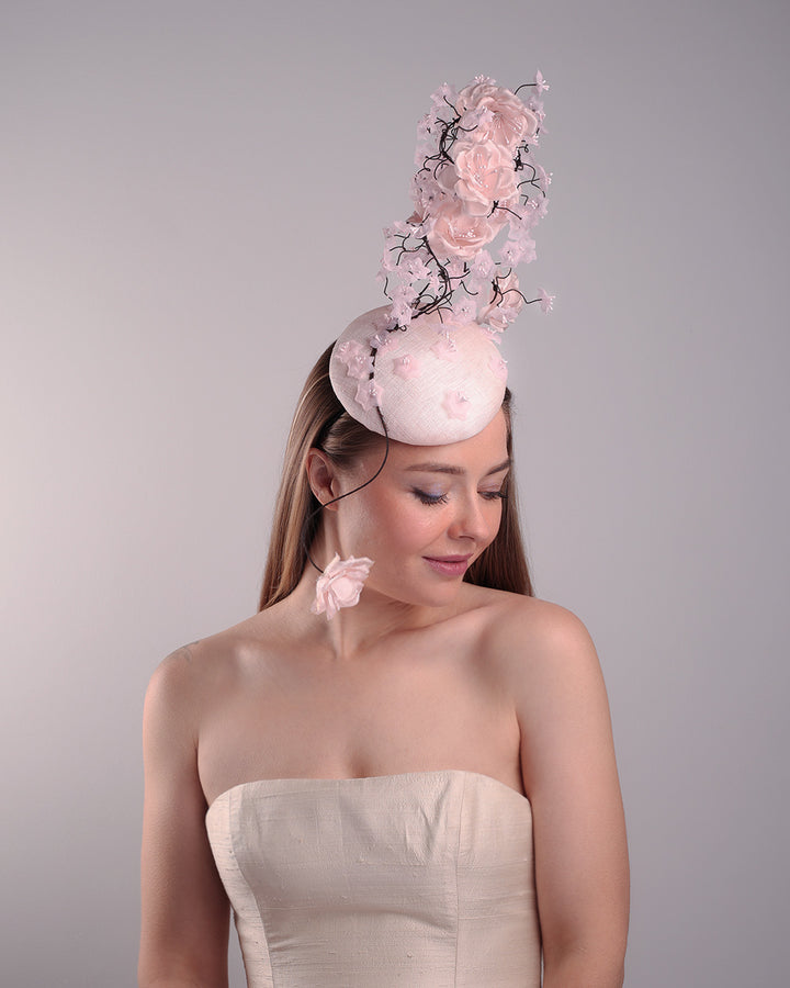 Model is showcasin the right angle of the button shaped pink fascinator with sinamay base decorated with tree like wire branches embellisehd with custom made pink silk flowers. The pink button fascinator is placed on the right front side of the face and the wire braces go up towards the left side of the button base while a single branch with one rose falls on the right side of the face.