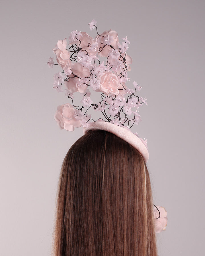 Model is showcasing the back of the button shaped pink fascinator with sinamay base decorated with tree like wire branches embellisehd with custom made pink silk flowers. The pink button fascinator is placed on the right front side of the face and the wire braces go up towards the left side of the button base while a single branch with one rose falls on the right side of the face.