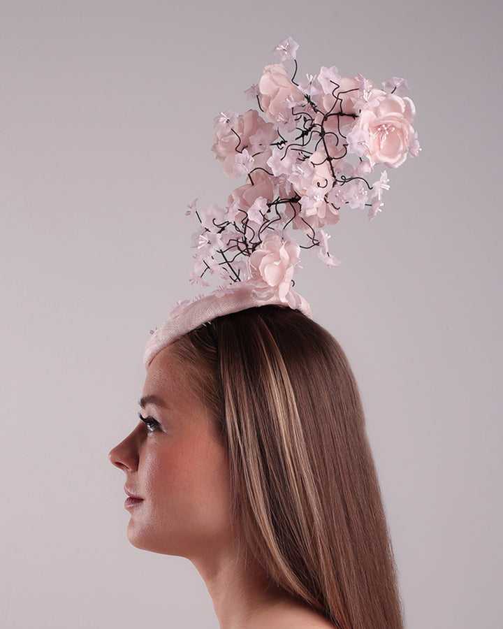 Model is showcasing the left side of the button shaped pink fascinator with sinamay base decorated with tree like wire branches embellisehd with custom made pink silk flowers. The pink button fascinator is placed on the right front side of the face and the wire braces go up towards the left side of the button base while a single branch with one rose falls on the right side of the face.
